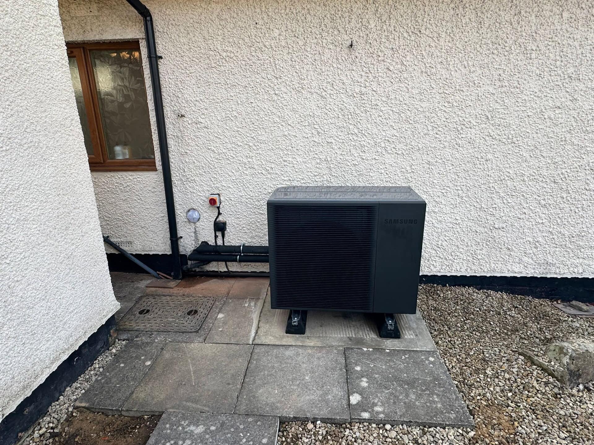 A black Samsung Air Source Heat Pump Unit against a white washed wall.