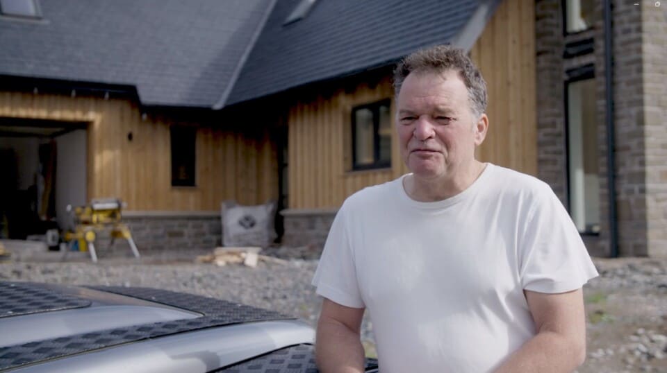 A man stands on a building site, outside a large detached house that is almost built.