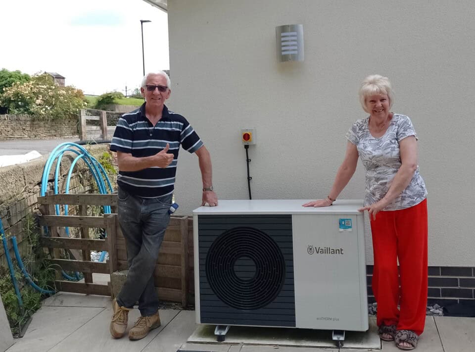 Sue and Mike Housley stood next to thier 5kW Vaillant Air Source Heat Pump outdoor unit. Sue is wearing a white top with a black pattern on it with red trousers and fluffy slippers, she is smiling and leaning on the heat pump. Mike is on the left of the heat pump and is wearing a blue and white striped top and dark blue jeans and work boots. He is smiling and giving a thumbs up.