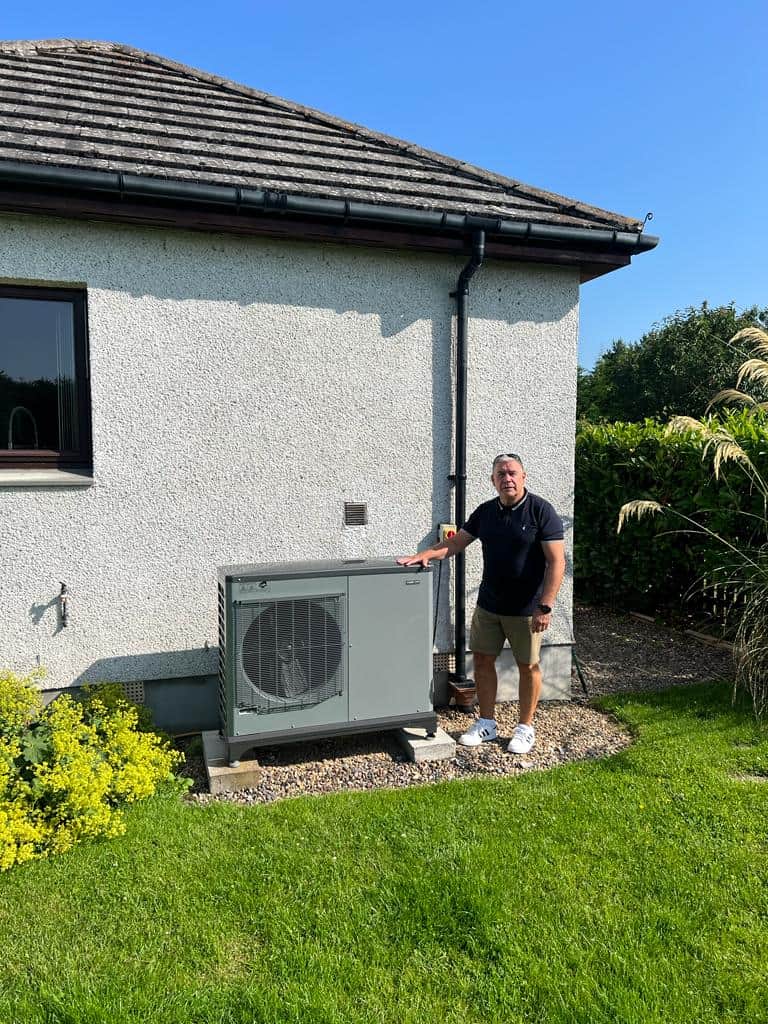 Picture of a man stood next to an Air Source Heat Pump outside his bungalow in the sunshine.