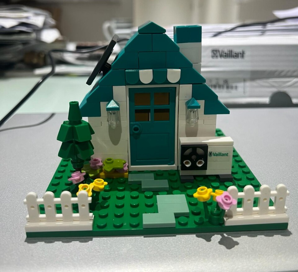 A 'lego' house with a green roof and white walls, sat on 'lego' grass with a white fence and flowers around it. Next to the house is a 'lego' air source heat pump' - all made with Lego.