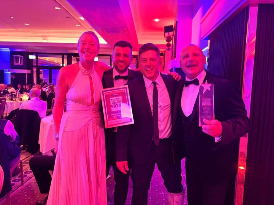 Emma Bohan, Managing Director of IMS Heat Pumps, stands with Christian Holmes, Heating Engineer, Lee Brown, Operations Director, and Dave Lakin, Senior Heating Engineer, at the National ACR & Heat Pump Awards 2026. Dressed in formal evening wear, the team are smiling and celebrating after winning Domestic Air to Water Heat Pump Project of the Year, proudly displaying their winner's certificate and trophy. The awards ceremony takes place in a brightly lit ballroom with fellow industry guests seated in the background.