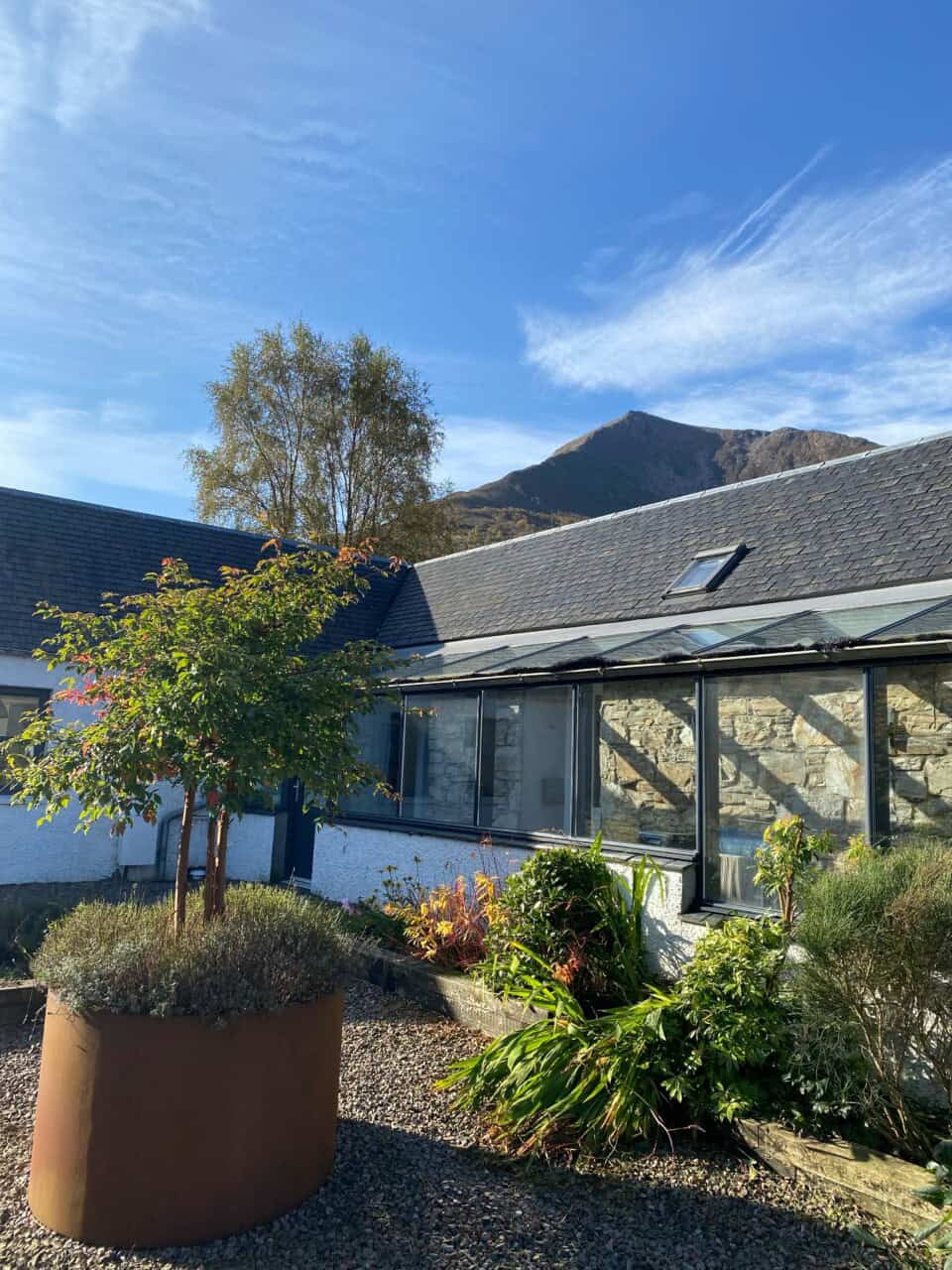 Close-up of the Jacksons' home courtyard with a modern extension featuring glass panels, a potted tree in the foreground, and stunning Highland peaks visible in the background.